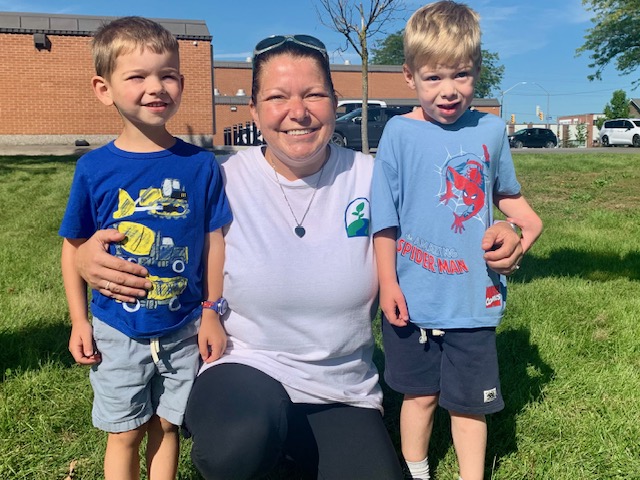 Smiling woman crouching between two young boys on the grass outside Rivergate Academy