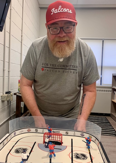 Pete Montana smiling over a tabletop hockey game, wearing a Falcons cap and Falcon Hockey shirt