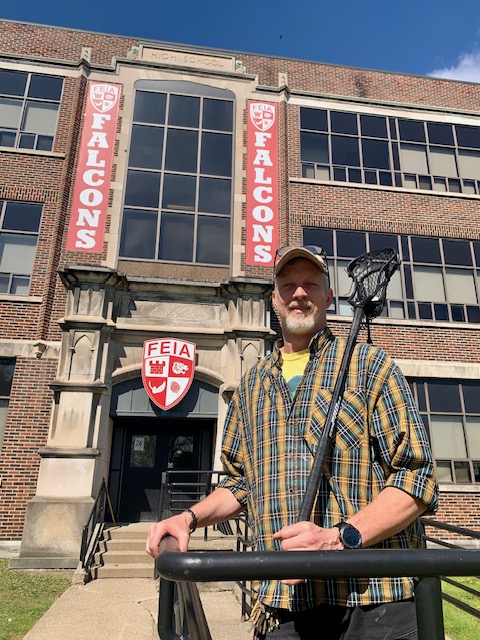 Jason Vehrs holding a lacrosse stick in front of the FEIA school entrance with Falcons banners