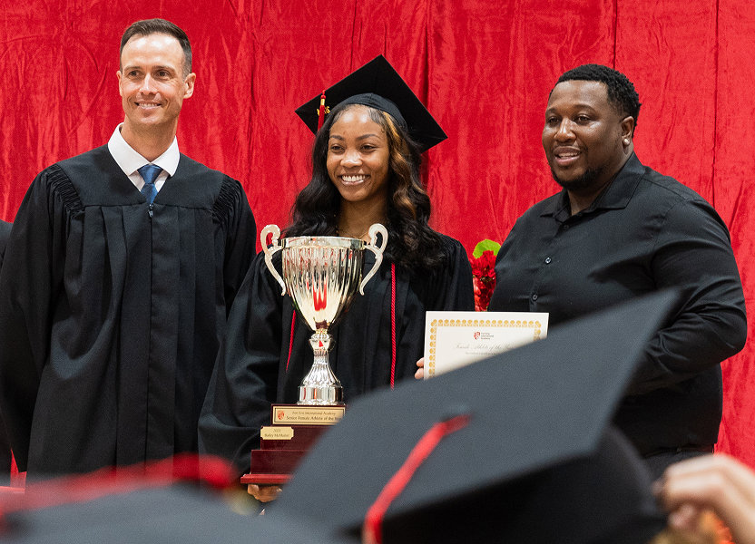 FEIA graduate in cap and gown holding a trophy beside two men at a graduation ceremony