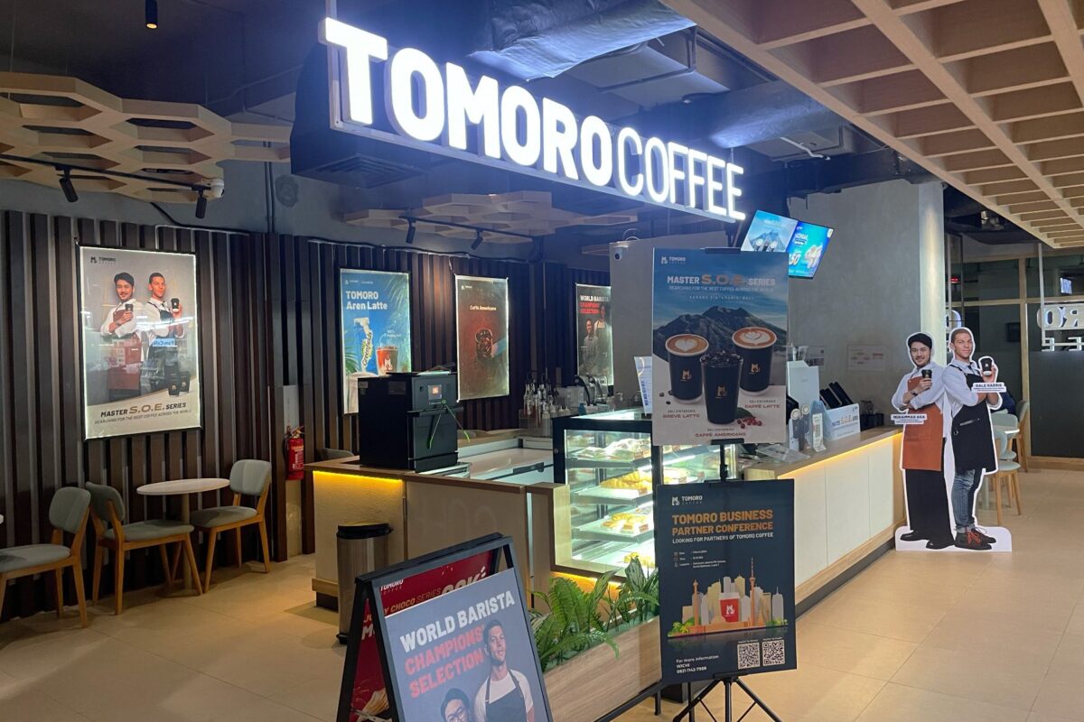 Suasana kedai TOMORO Coffee dengan papan menu dan poster promosi di area kasir, menampilkan konsep kedai kopi modern dan digital.
