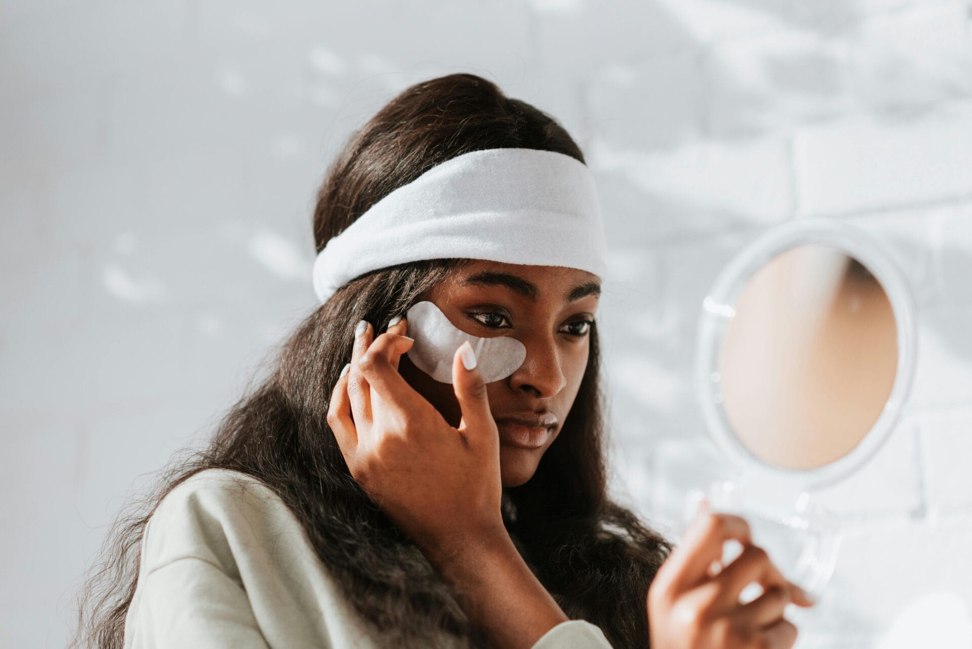 A person with long hair wears a white headband and applies an under-eye patch rich in antioxidants while looking into a handheld mirror, focusing on their skincare routine.