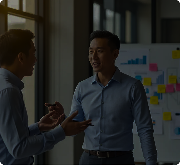 Two men in professional attire having a discussion in an office with charts and sticky notes on a board behind them.