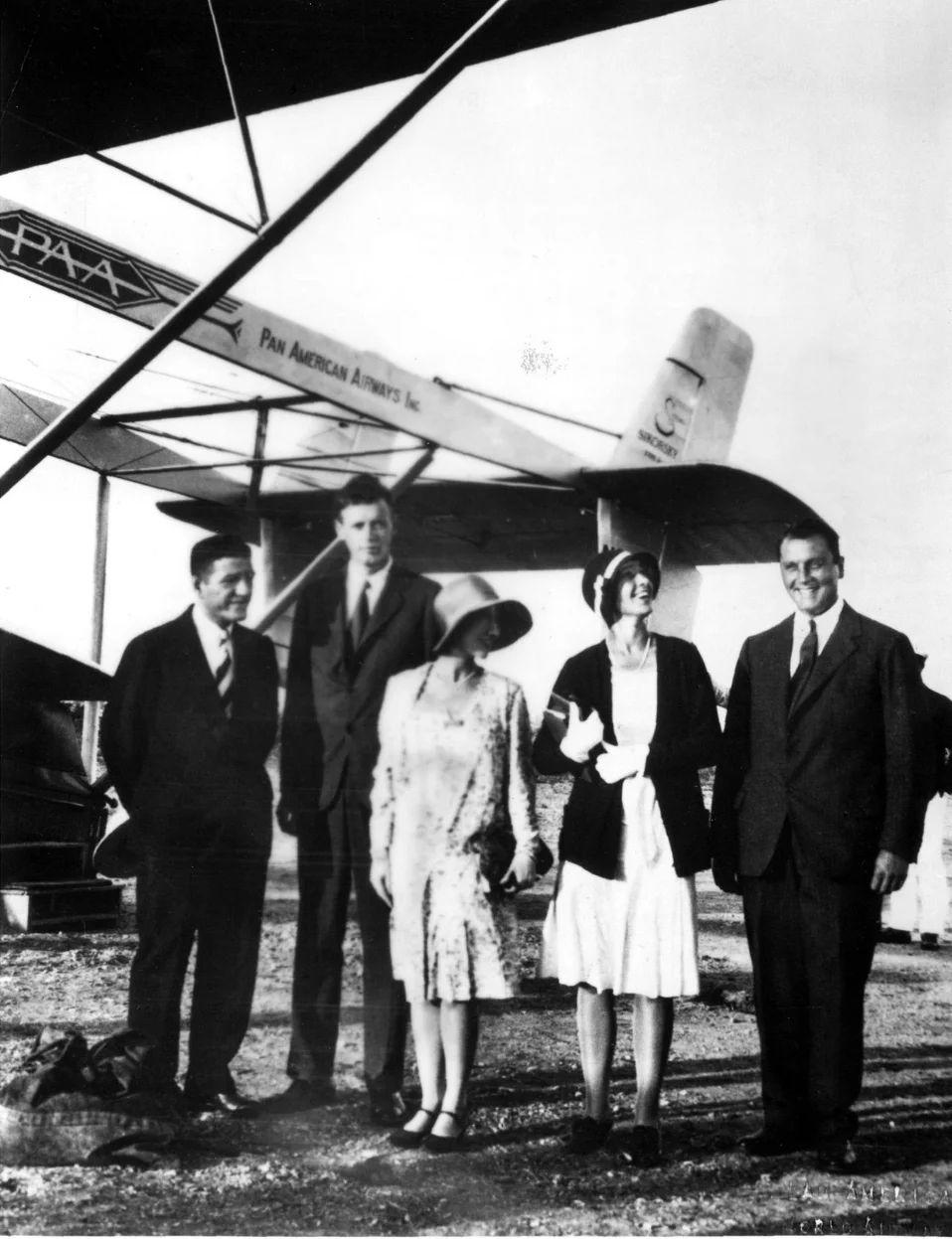 Black and white photo of three men in suits and two women in dresses and hats standing in front of a Pan American Airways airplane.