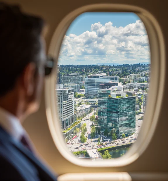 Man in suit looking out airplane window at a cityscape with modern buildings and cloudy sky.