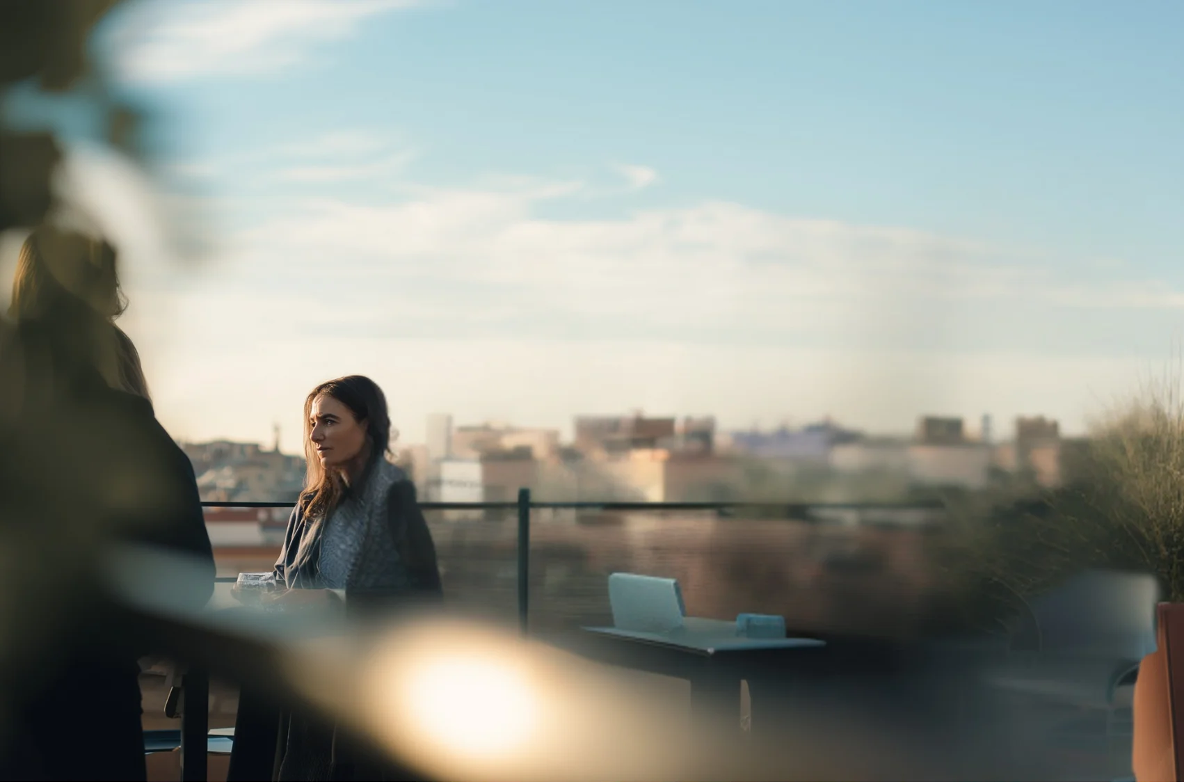 Two women having a conversation on a rooftop terrace with a cityscape in the background during daylight.