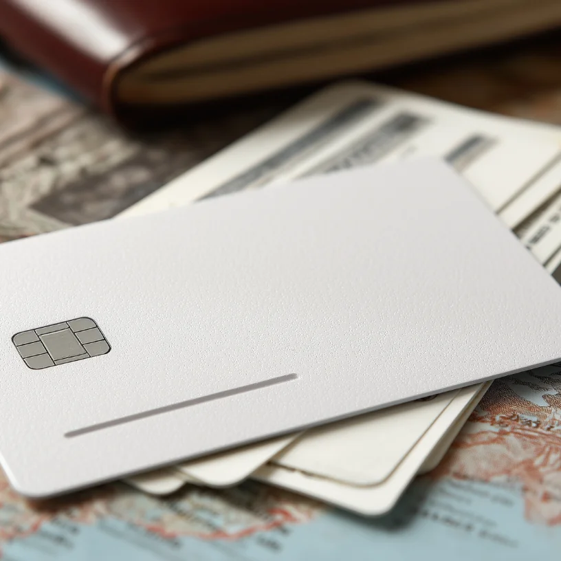 Close-up of a blank white credit card with a chip on top of several other cards and a brown leather wallet on a map background.