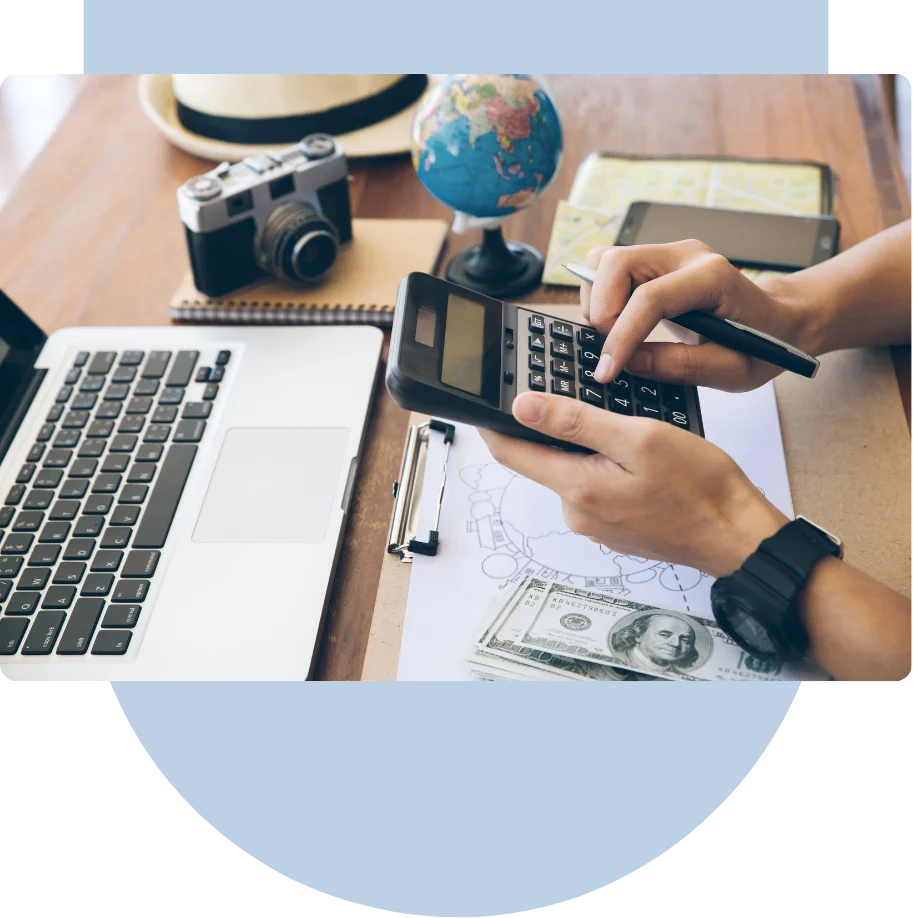 Person using a calculator with a laptop, dollar bills, camera, globe, and a map on a wooden desk.