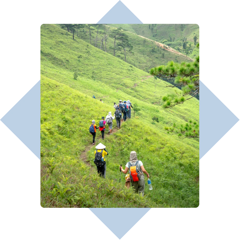 Group of hikers walking in single file along a narrow dirt trail through lush green hills.