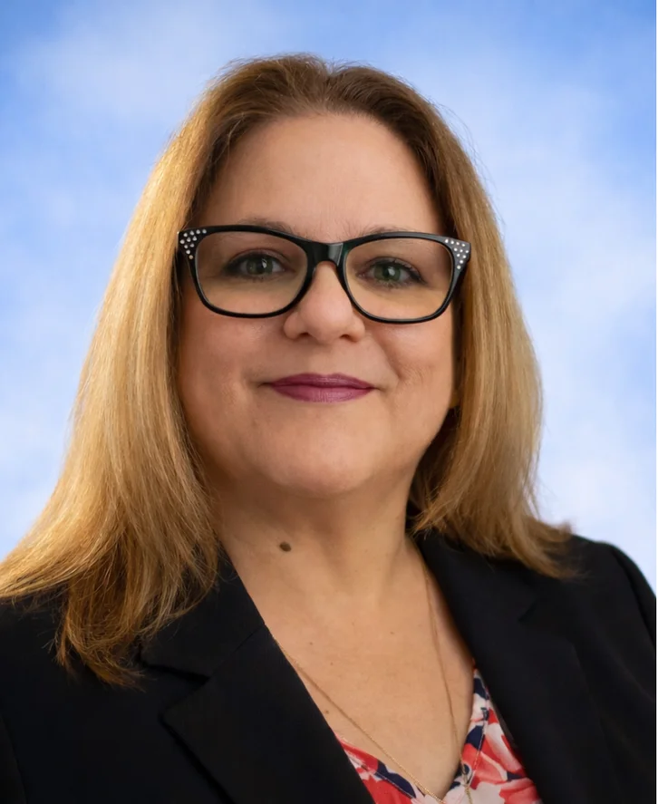 Smiling woman with shoulder-length light brown hair, black framed glasses, and wearing a black blazer over a floral top against a blue sky background.