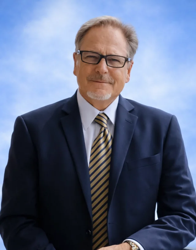 Man with glasses wearing a dark suit and striped tie standing on a balcony with a scenic outdoor background.