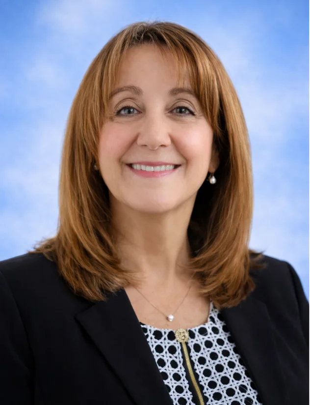 Smiling woman with shoulder-length light brown hair wearing a patterned black and white top and pearl earrings.