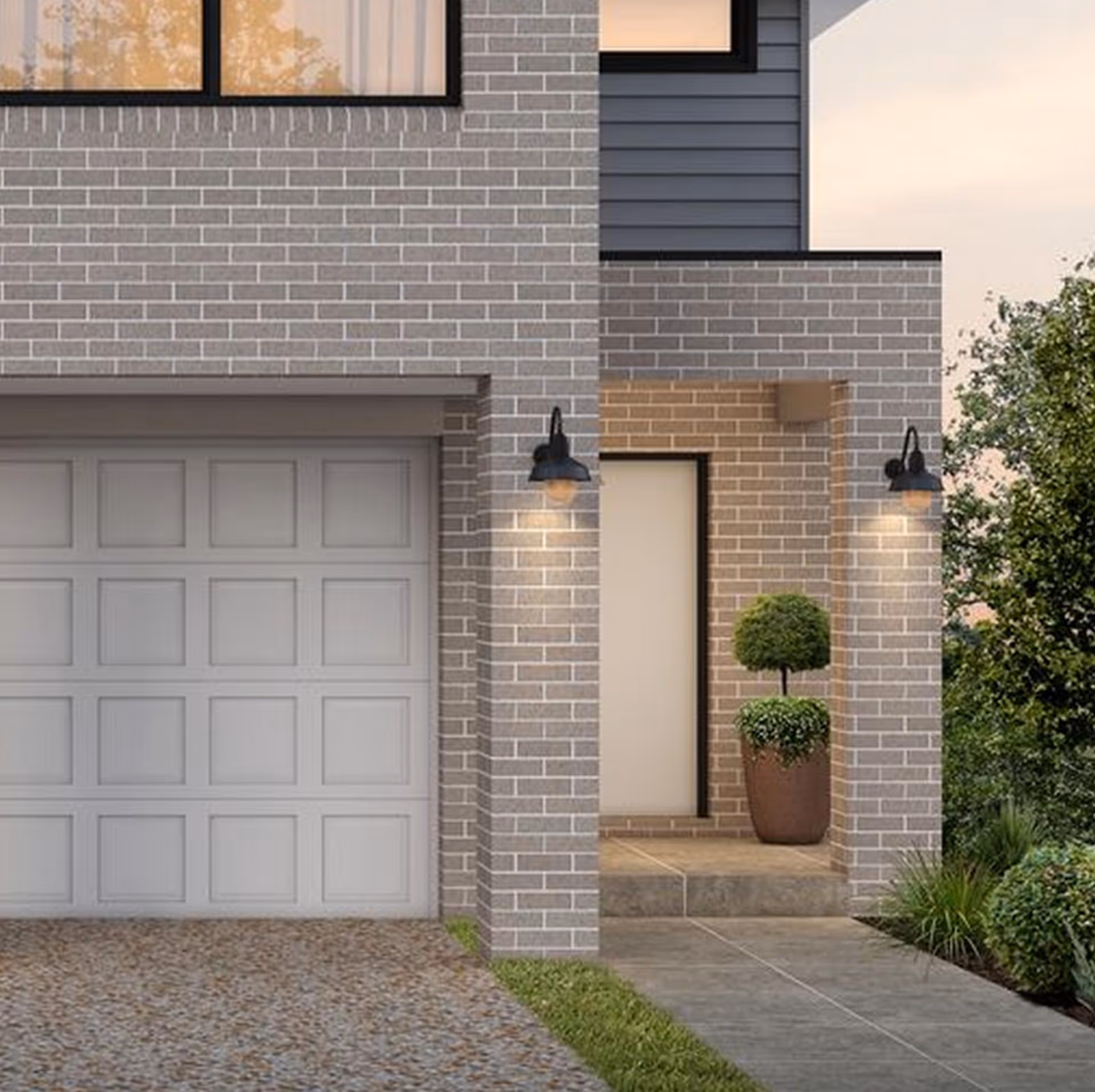Modern house facade with light gray brick walls, a white garage door, illuminated porch with twin lamps, and potted topiary plant by the entrance.
