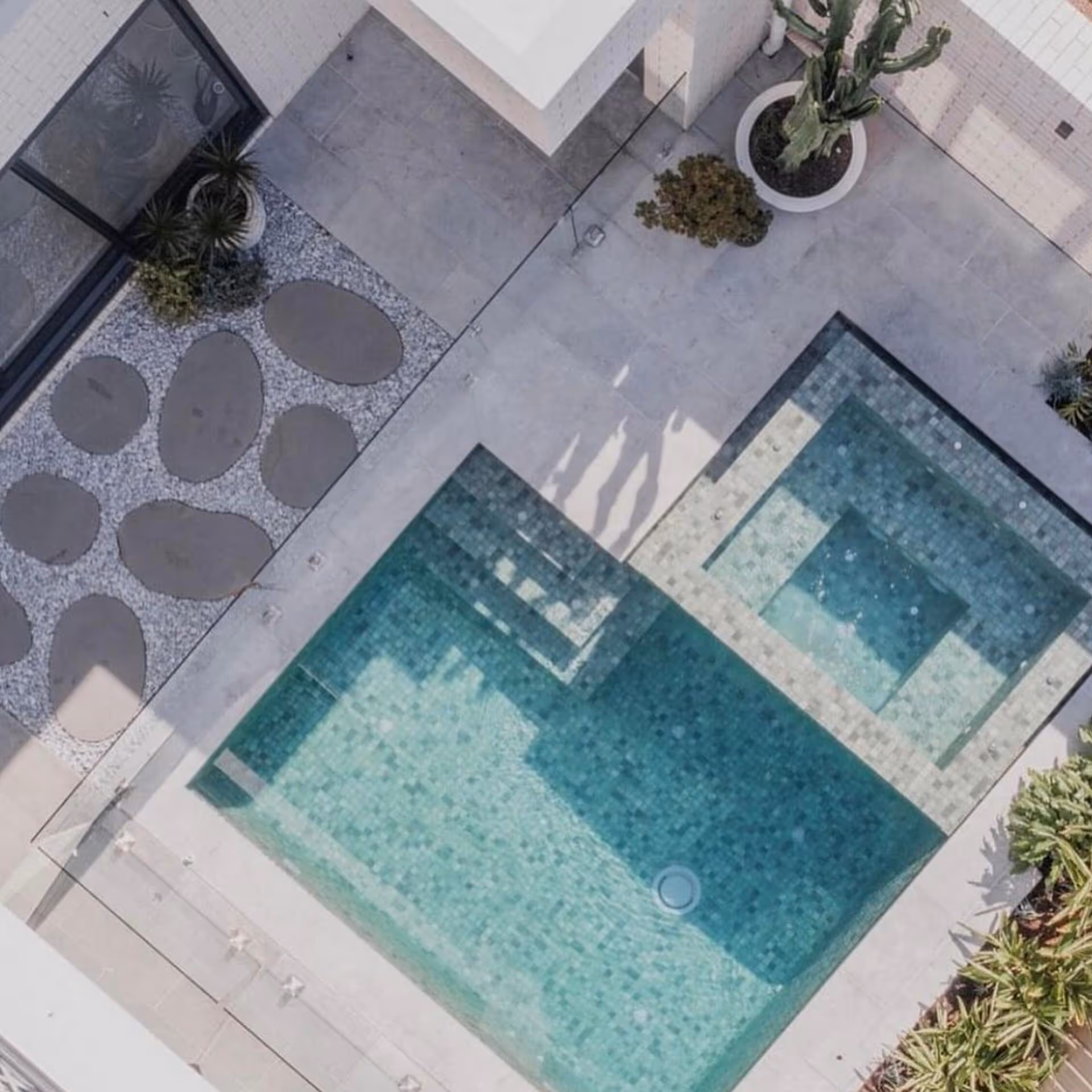 Aerial view of a modern swimming pool with geometric designs, surrounded by a concrete patio and decorative plants. Shadows of two figures are visible.
