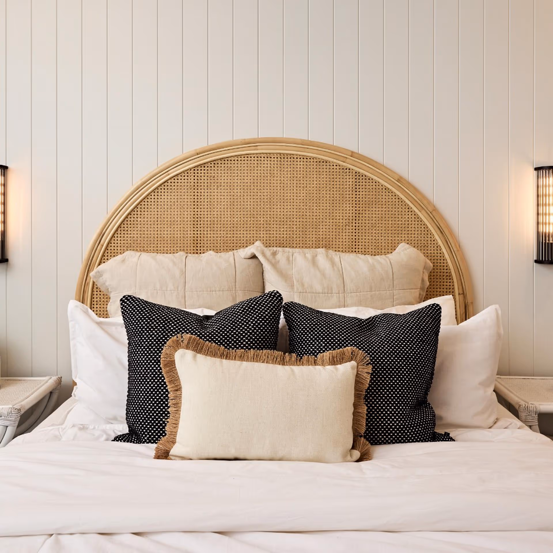 A cozy bed with a wicker headboard, adorned with beige and black patterned pillows. The soft lighting creates a warm, inviting atmosphere.