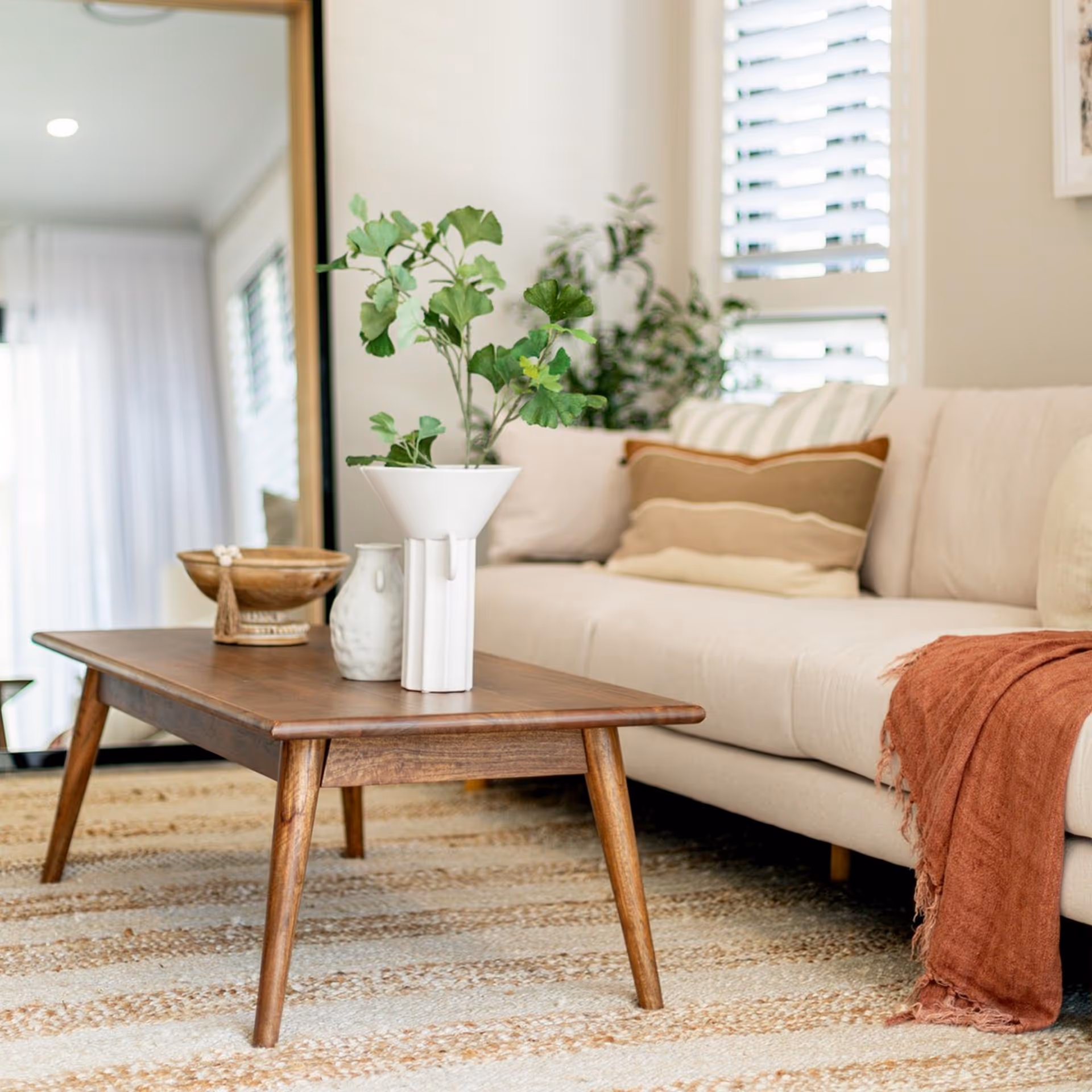 Cozy living room with a beige sofa adorned with cushions and a rust-colored throw. A wooden coffee table with a white vase and green plant sits on a textured rug.