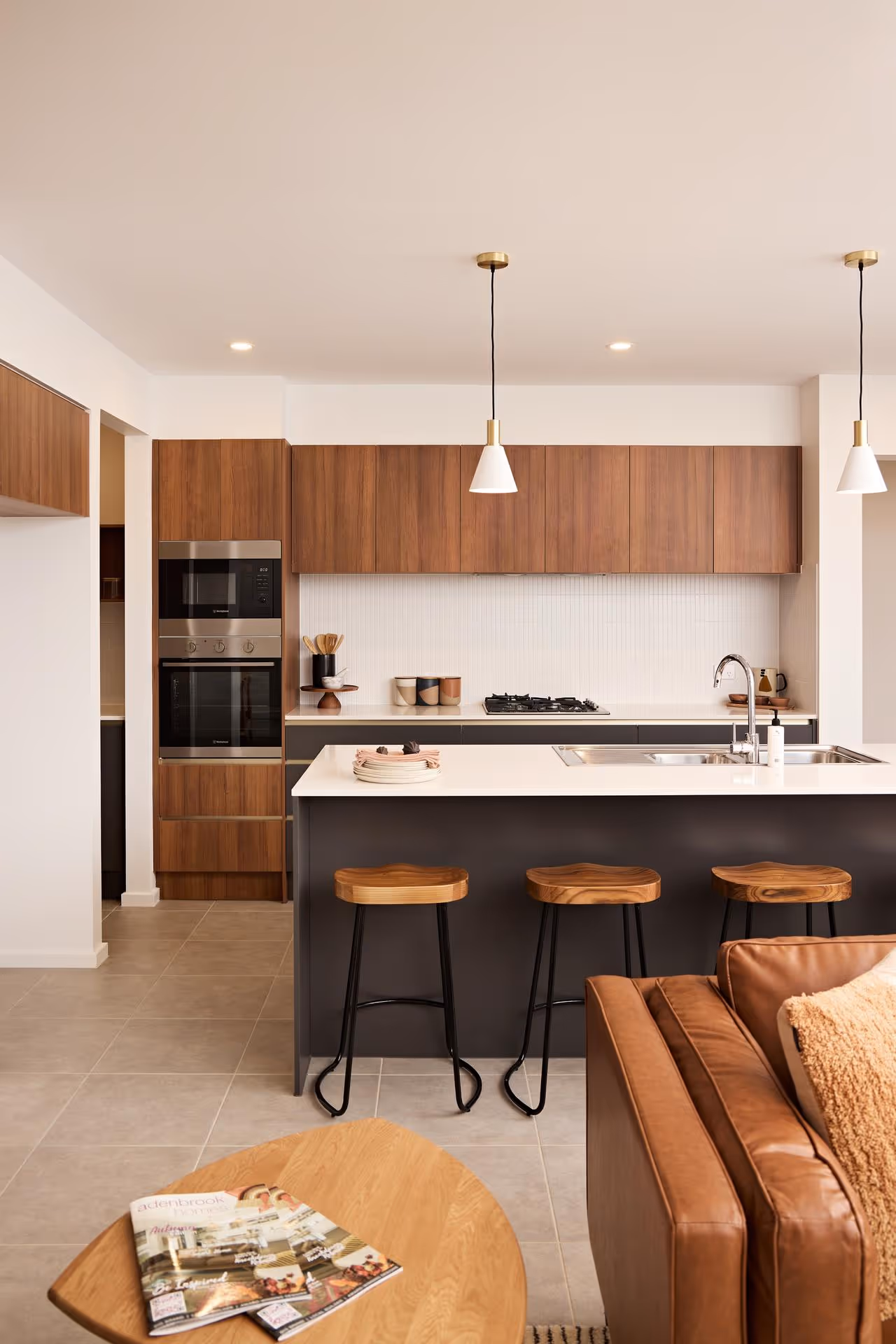 Modern kitchen with a warm tone, featuring wood cabinets, a white countertop, pendant lights, and black barstools. A cozy vibe with a brown leather sofa.