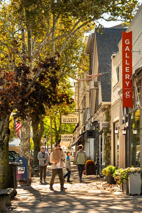 People walk along a sidewalk lined with shops, trees, and signs for a market, liquor store, and gallery on a sunny day.
