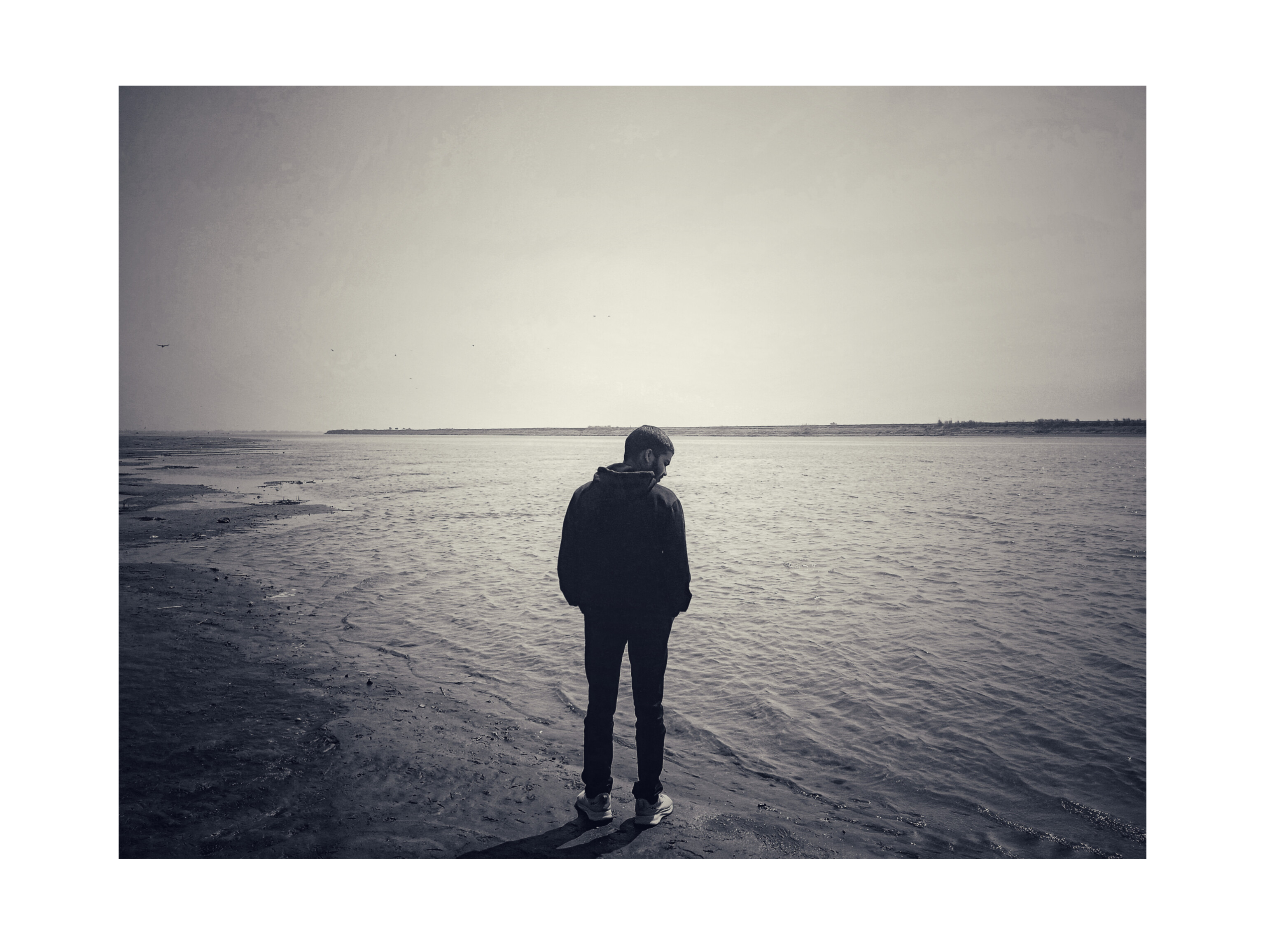 Person standing alone on a shore, looking down at the water on a cloudy day.