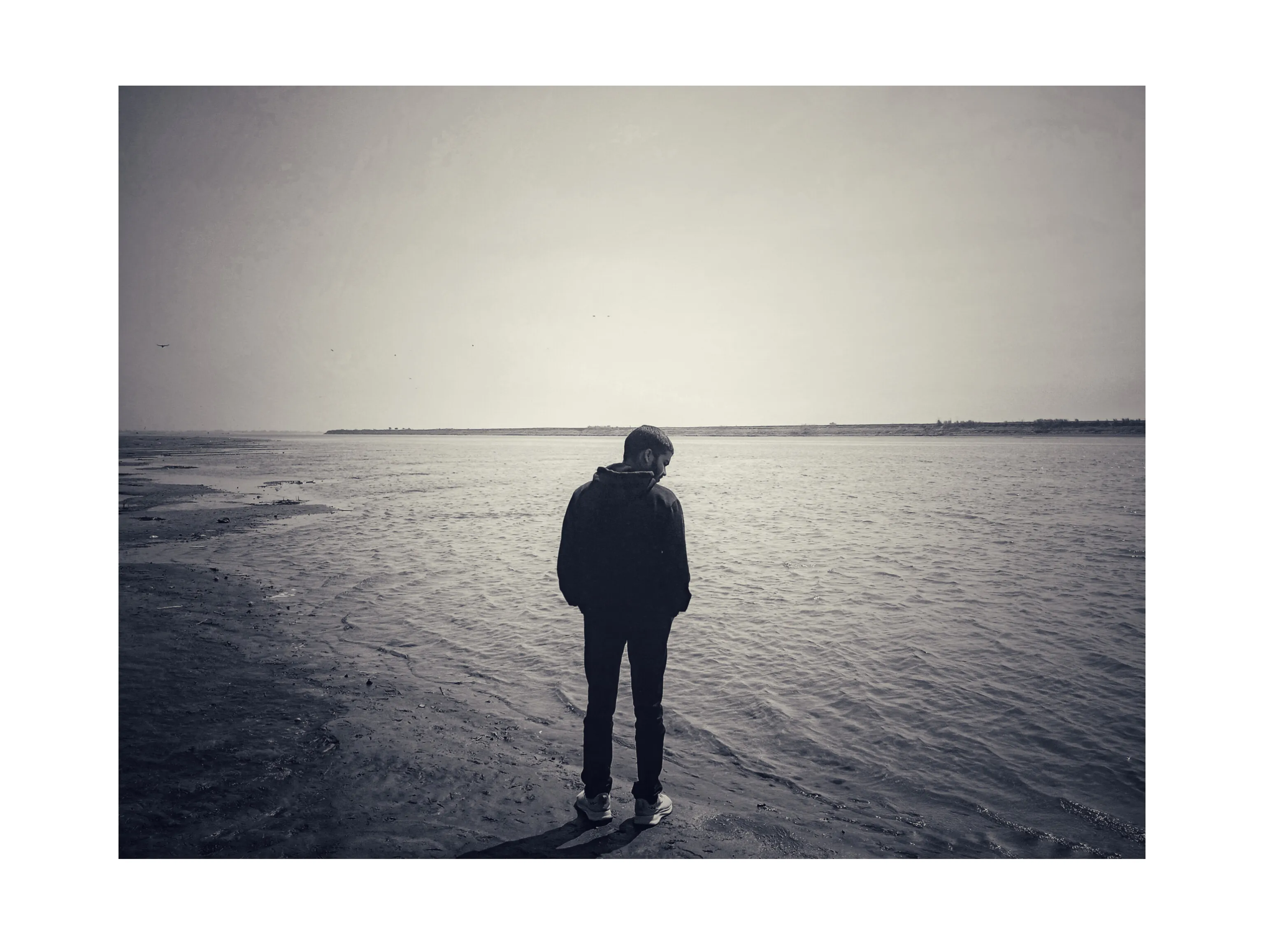 Person standing alone on a shore, looking down at the water on a cloudy day.