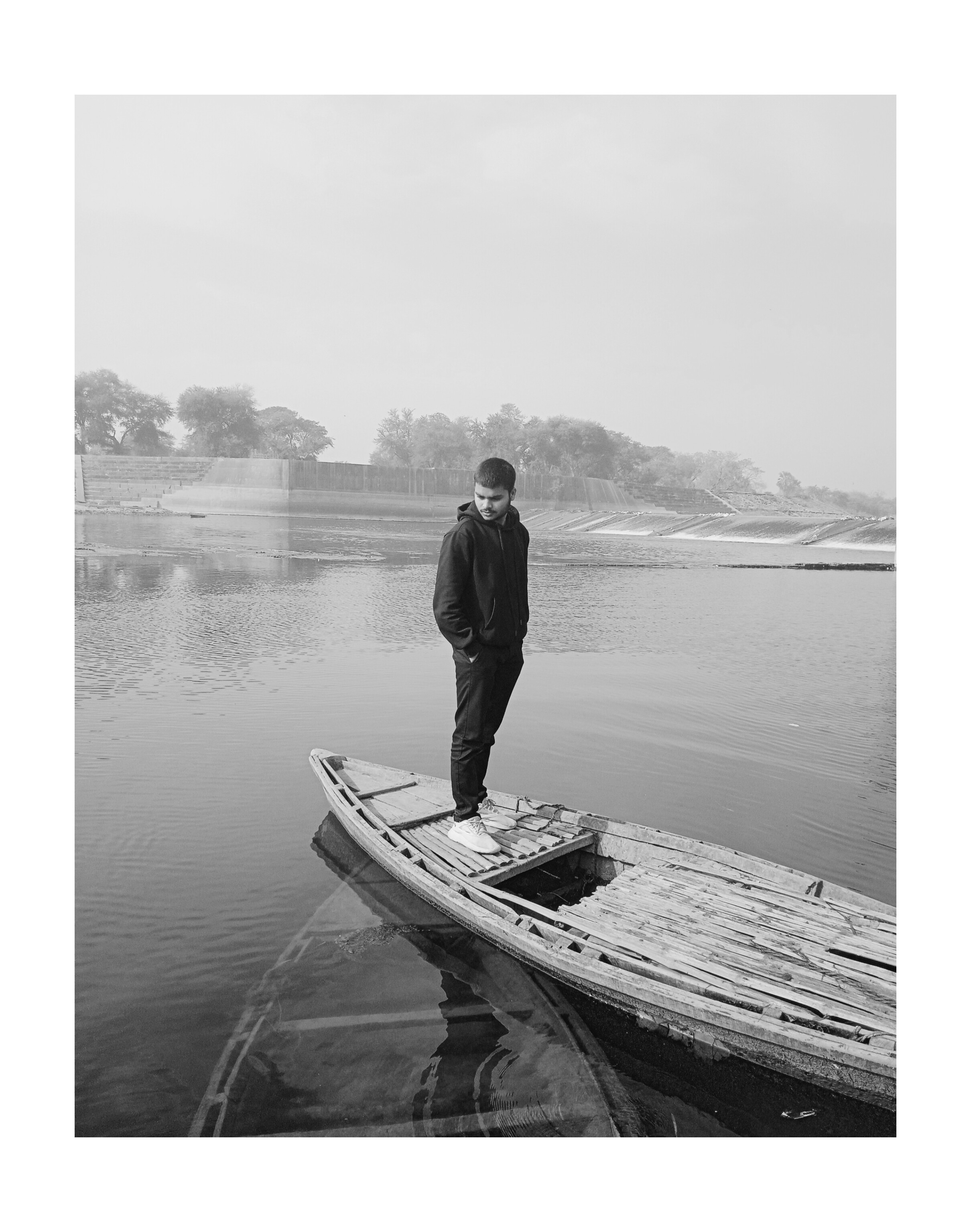 Man in a hoodie standing on an old wooden boat floating on calm water near a shoreline with trees and a concrete embankment.