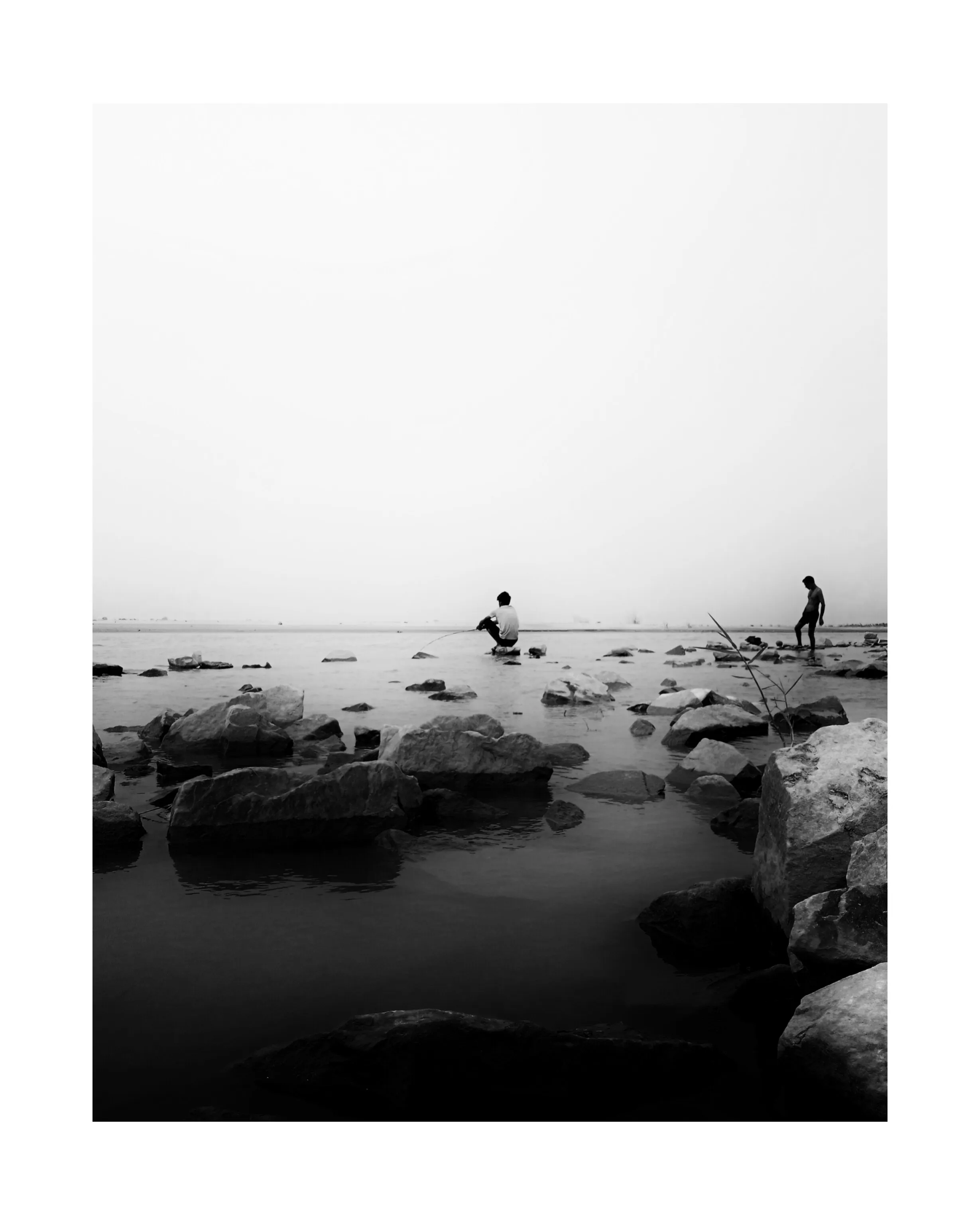 Black and white photo of two people fishing among rocks in calm shallow water under a bright sky.