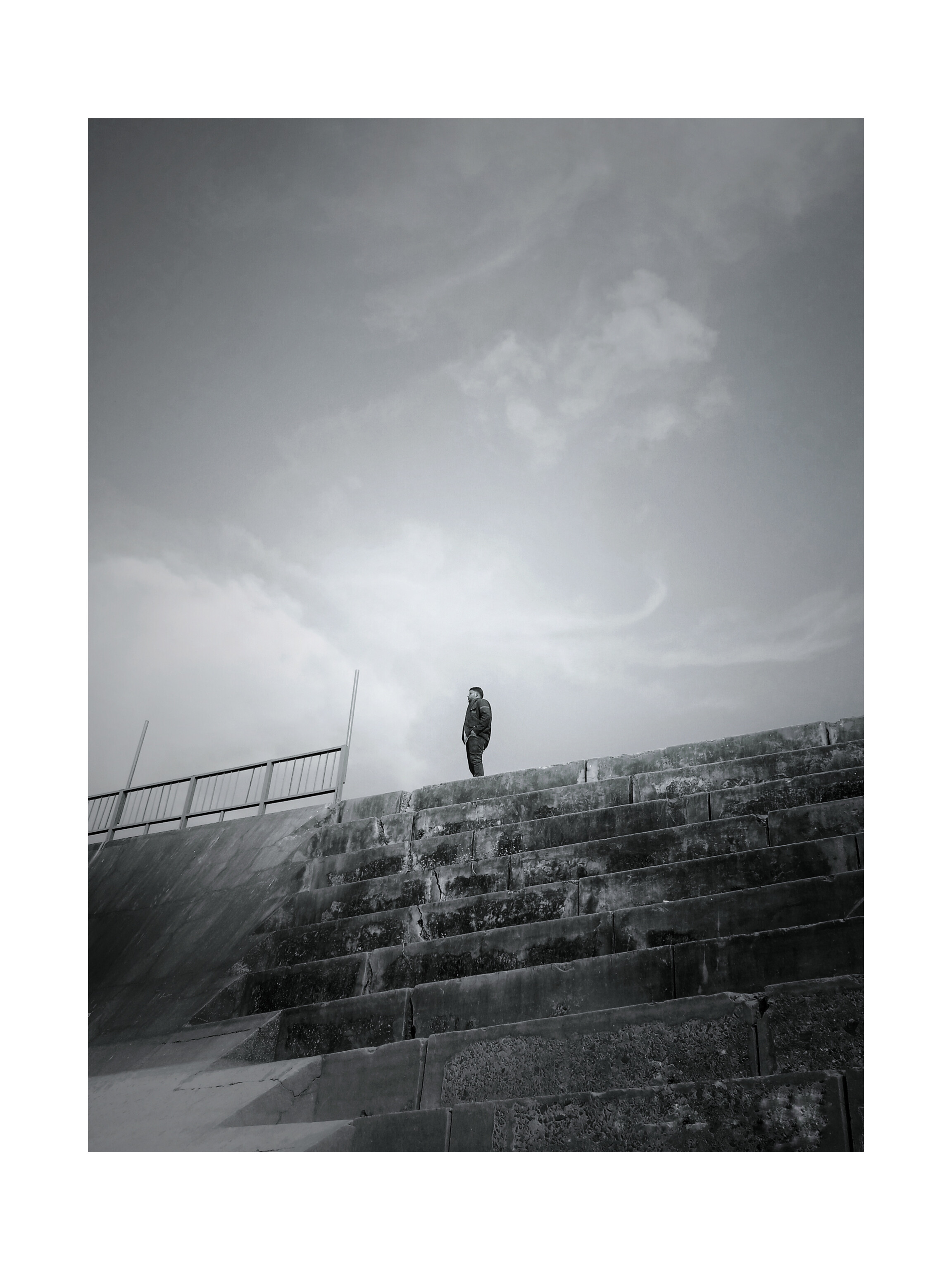 Black and white photo of a person standing alone atop weathered concrete steps under a cloudy sky.