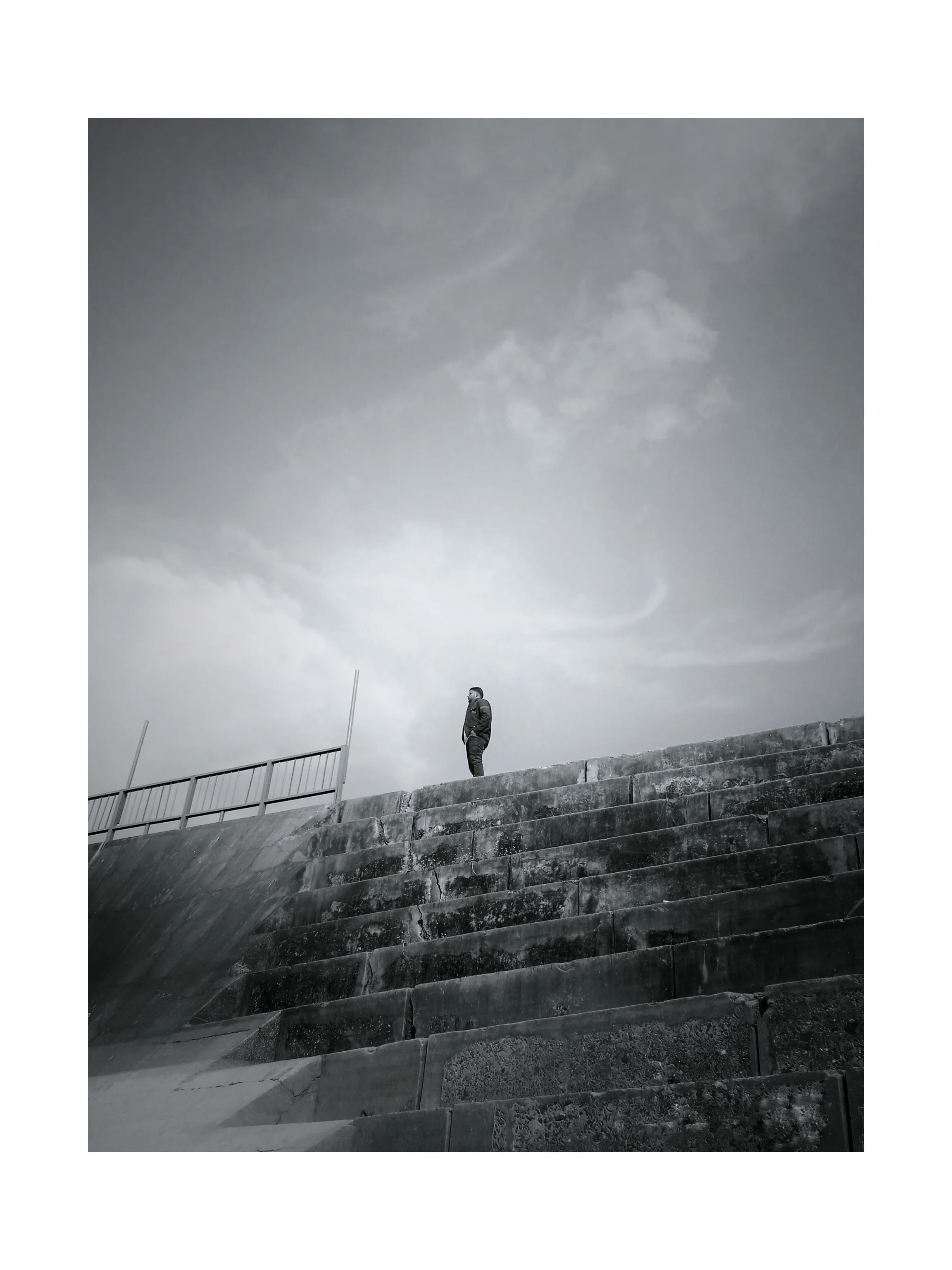 Black and white photo of a person standing alone atop weathered concrete steps under a cloudy sky.