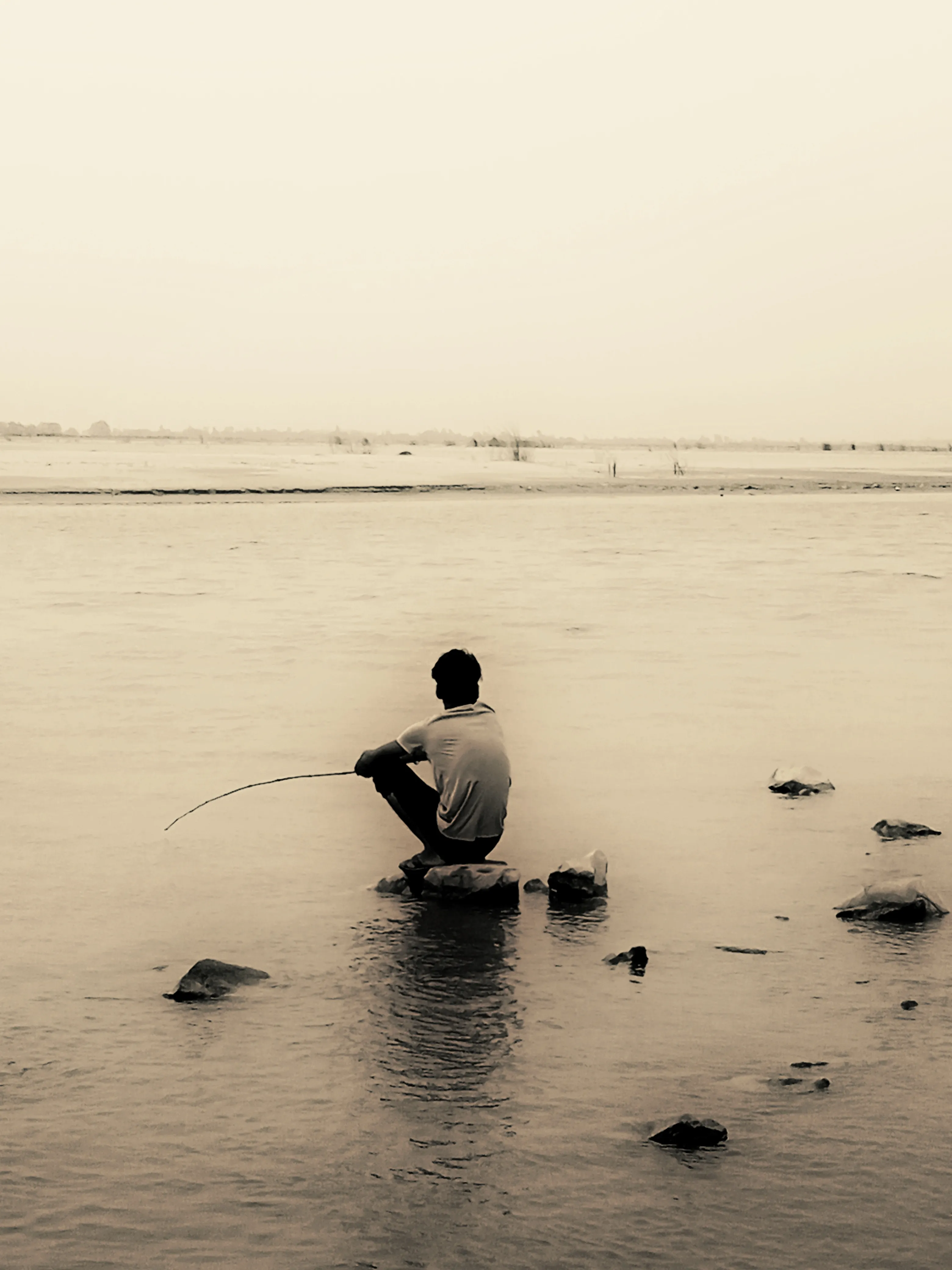 Person sitting on a rock in a body of water, fishing with a long, thin rod.