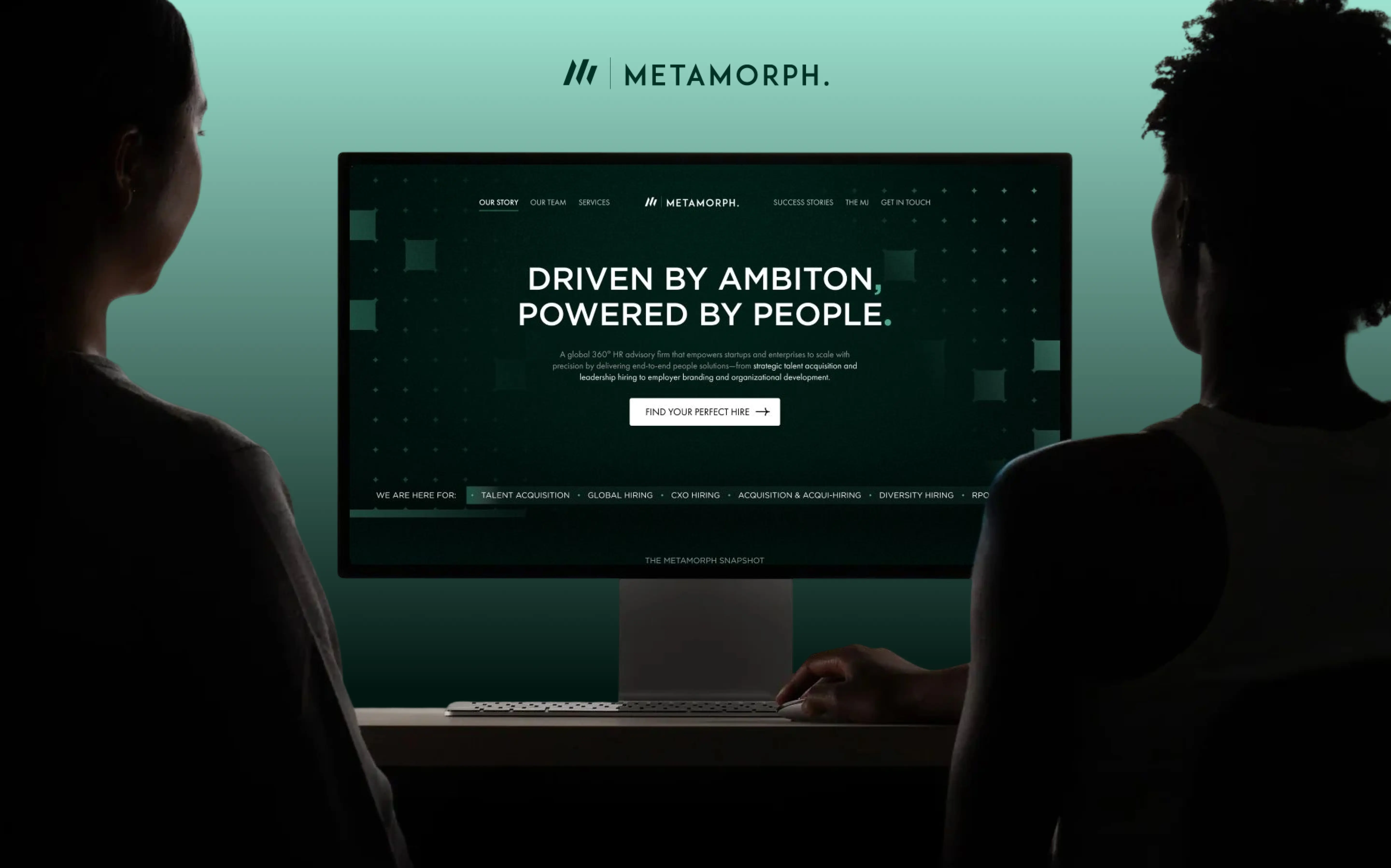 Two people viewing a computer screen showing Metamorph's website with the slogan 'Driven by ambition, powered by people.'