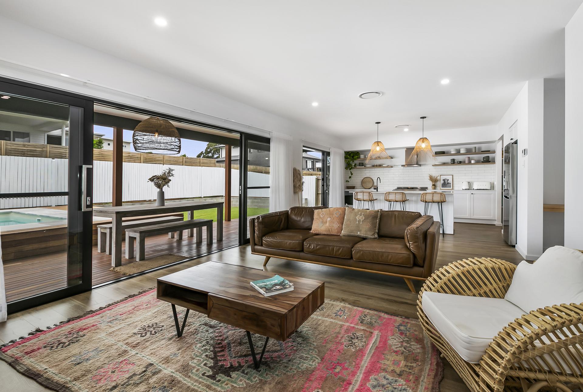 Modern living room with wooden floors, brown leather sofa, and wicker chairs. Glass doors open to a patio with a pool. Kitchen visible in the background.