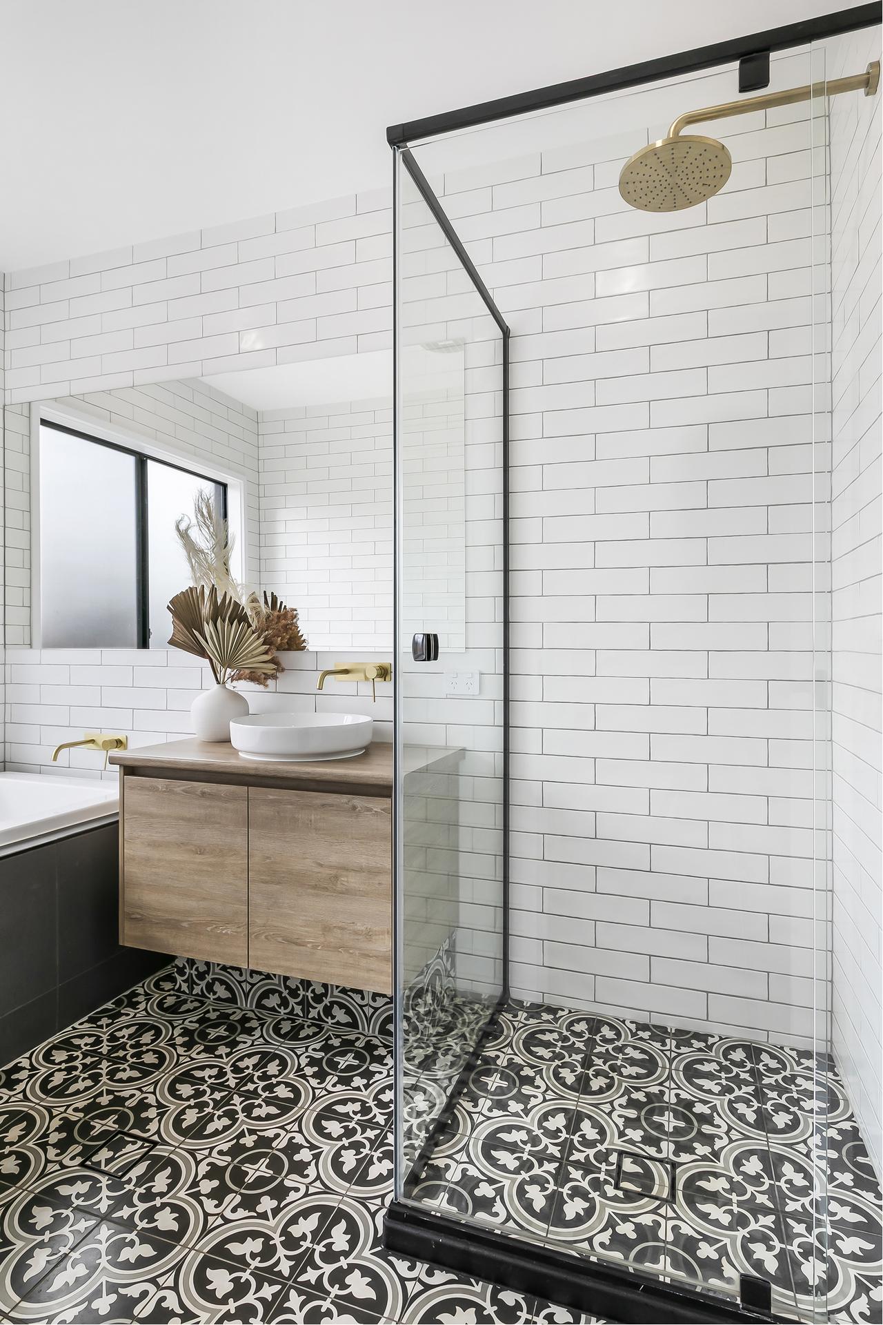 Modern bathroom with white subway tiles, glass shower, black patterned floor tiles. Wooden vanity with a round basin and gold fixtures, under soft light.