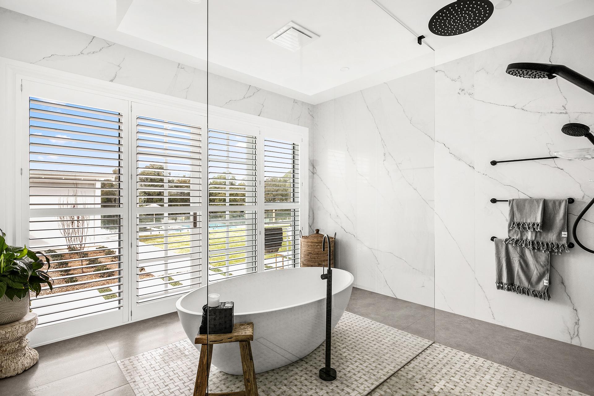 Luxurious bathroom with a freestanding oval bathtub, large rain showerhead, and floor-to-ceiling windows with shutters. Modern, serene, and elegant ambiance.