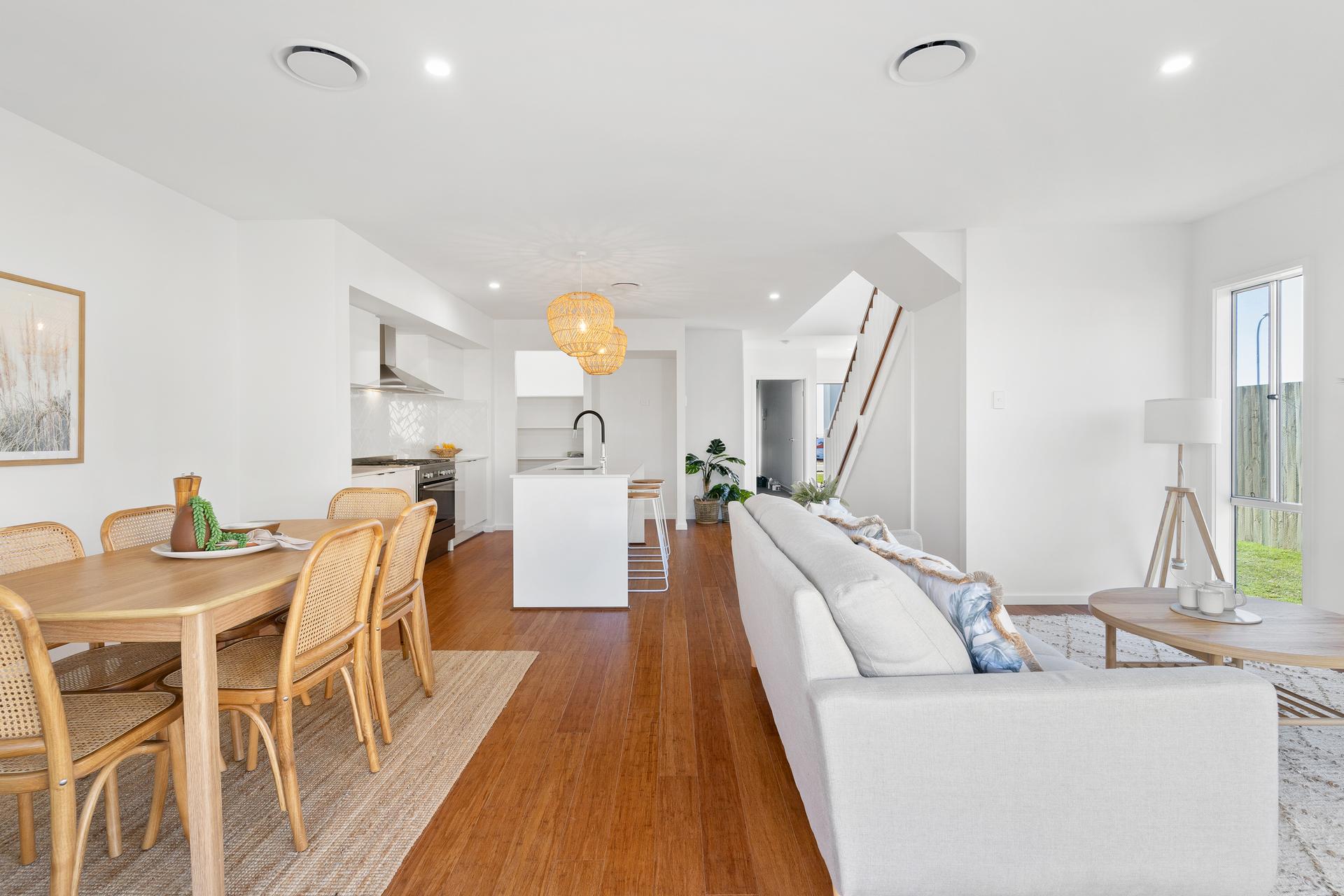 Modern open-plan interior with a bright dining and living area. Features wooden floors, a light gray sofa, dining table, island kitchen, and large windows.