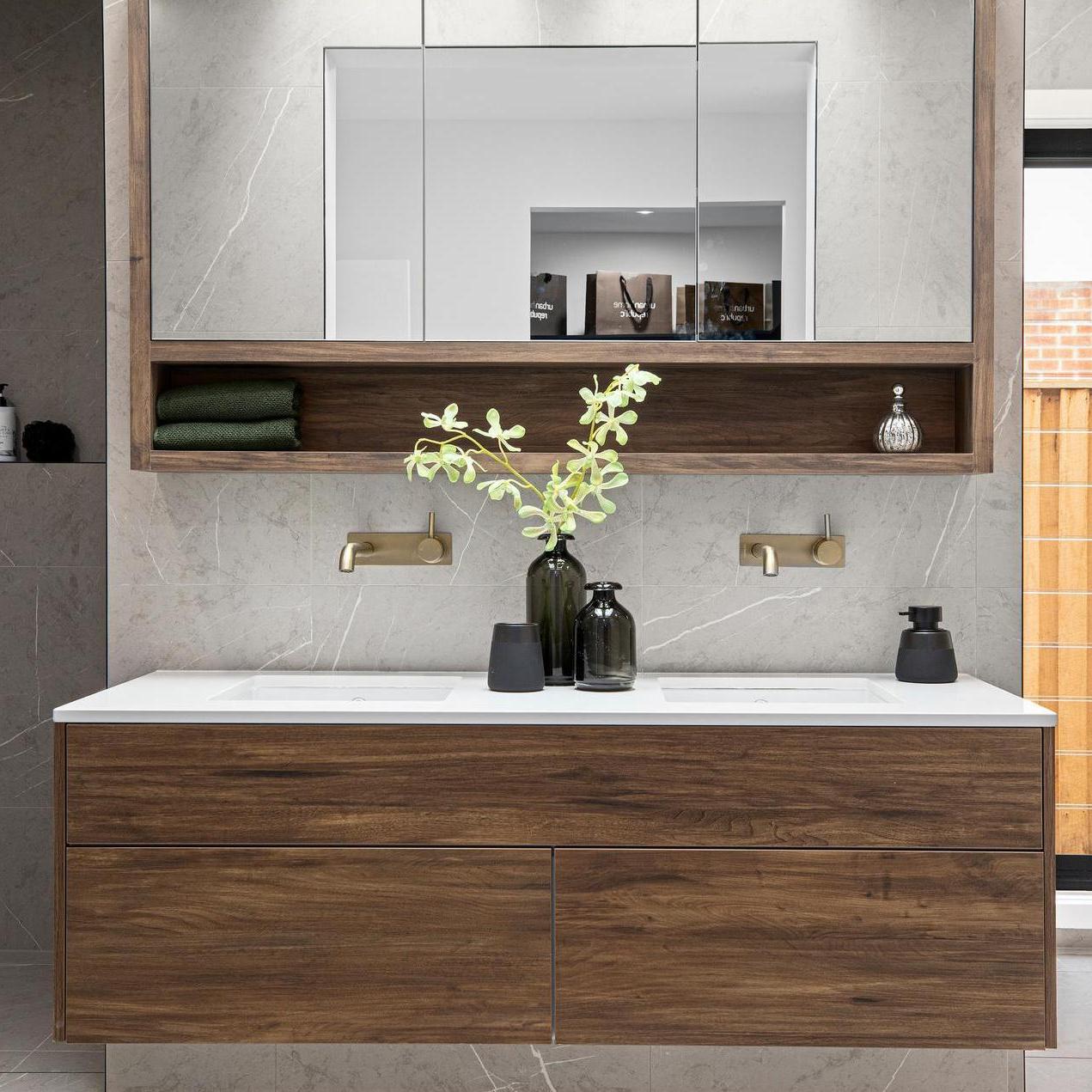 Modern bathroom with a sleek wood vanity, dual brass faucets, and marble backsplash. A small shelf holds a plant and decor, creating a calm, elegant tone.