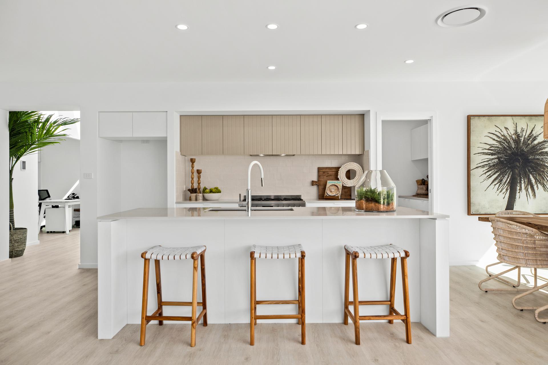 Modern kitchen with a white island and three wooden stools. Beige cabinets, minimalist decor, and a large palm artwork create a serene, airy atmosphere.
