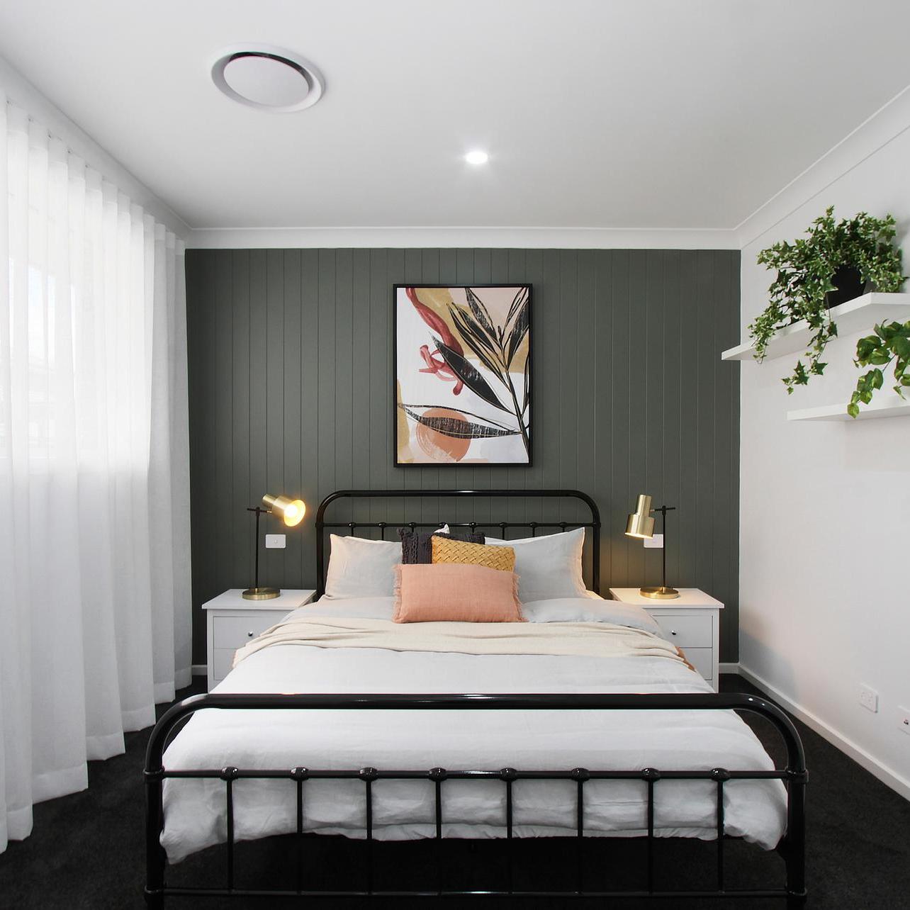 Minimalist bedroom with dark green accent wall, black metal bed, abstract artwork, white nightstands, warm lamps, white curtains, and green plants.