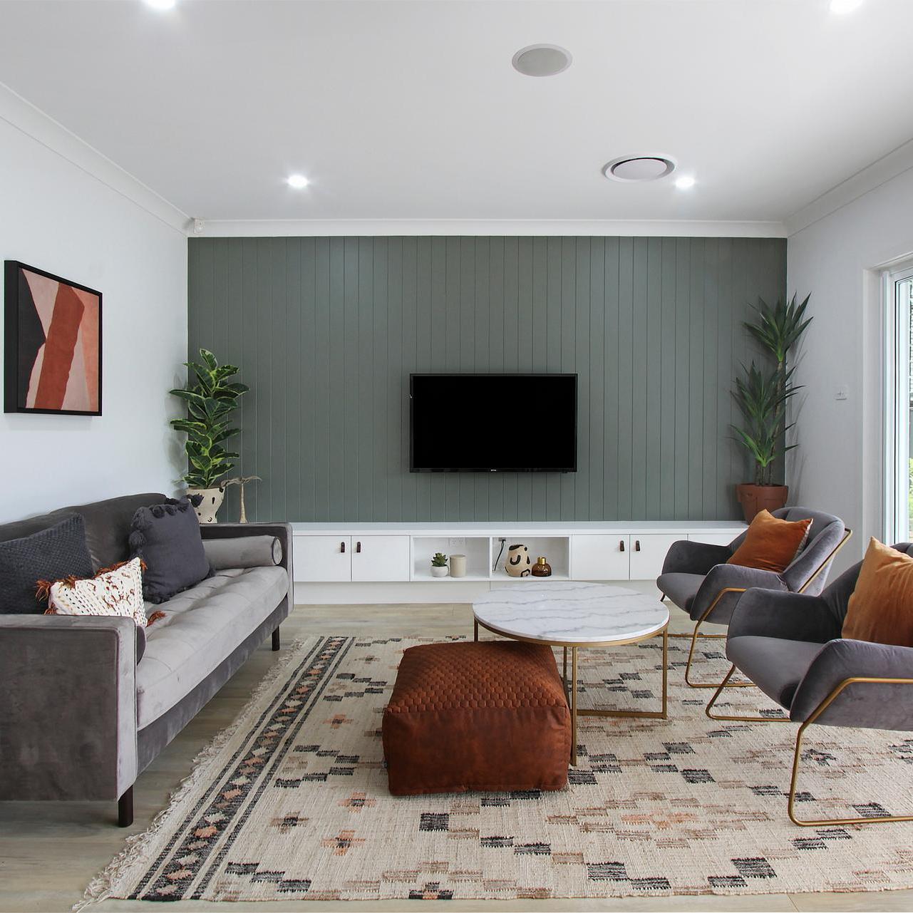 Modern living room with a gray sofa, abstract art, and a TV on a green accent wall. Features include stylish chairs, a marble coffee table, and a patterned rug.