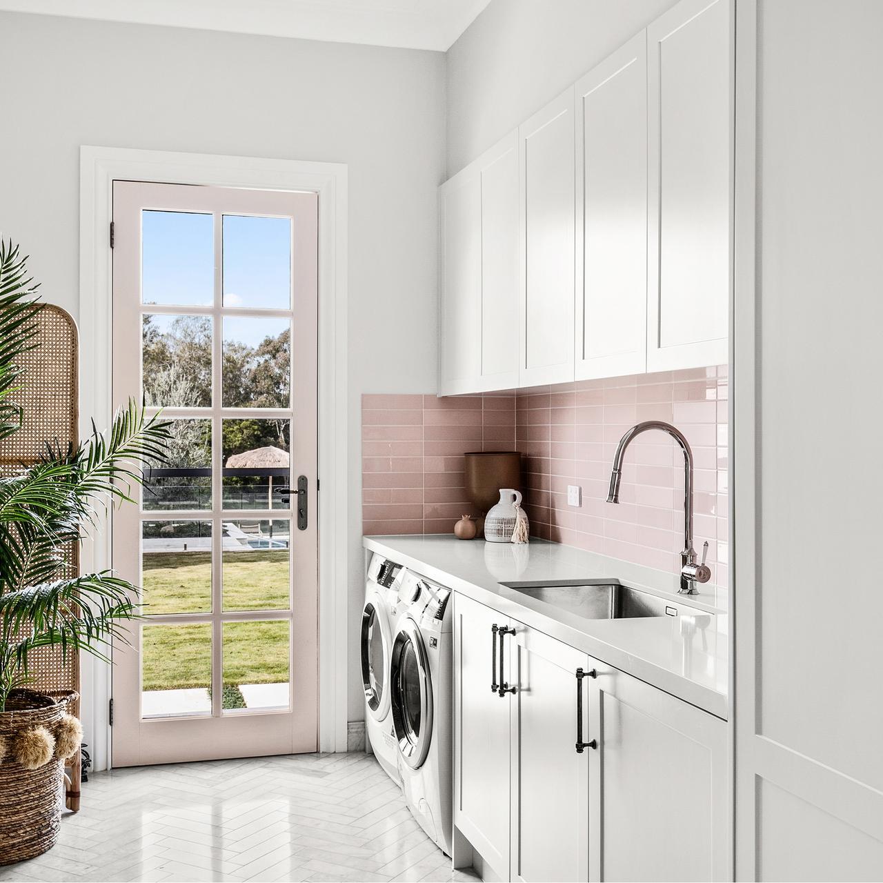 Bright laundry room with pink-tiled backsplash and white cabinets. A washer and dryer sit under a counter beside a sink. A glass door opens to a sunny garden.