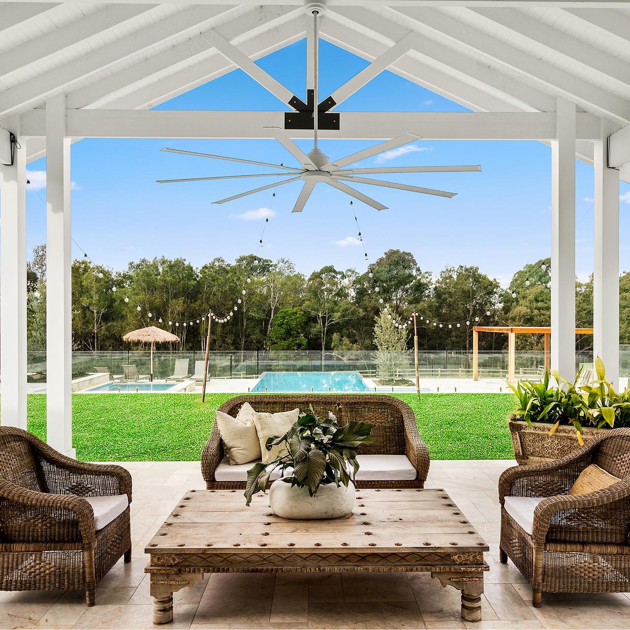 A cozy outdoor patio features wicker furniture with cushions, a large ceiling fan, and a wooden table with a plant. Beyond, a pool and lush trees create a serene atmosphere.
