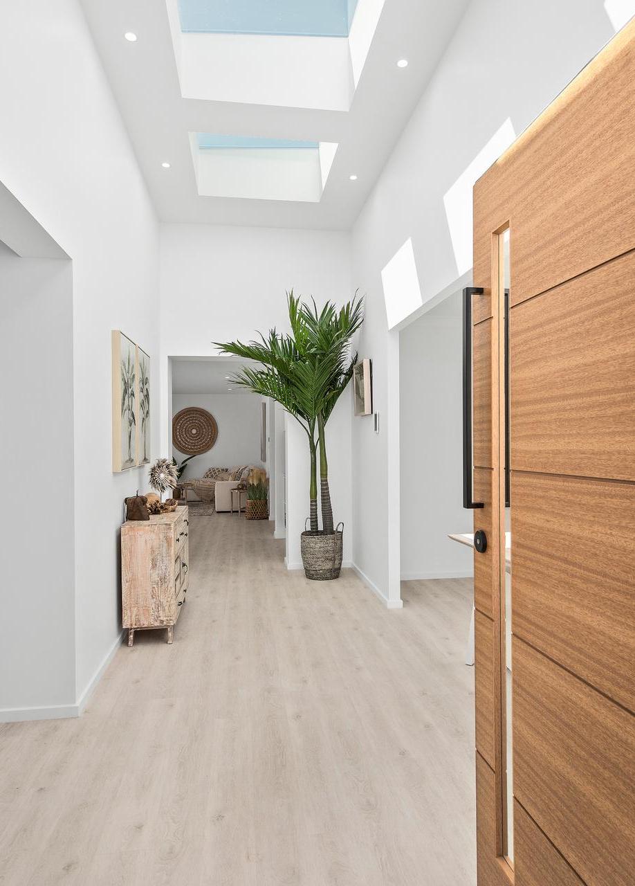 Modern hallway with light wood flooring, white walls, and ceiling skylights. A tall potted plant and rustic wooden cabinet add a natural touch.