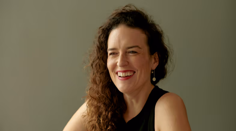Smiling woman with long curly hair wearing black sleeveless top and dangling earrings against a plain background.