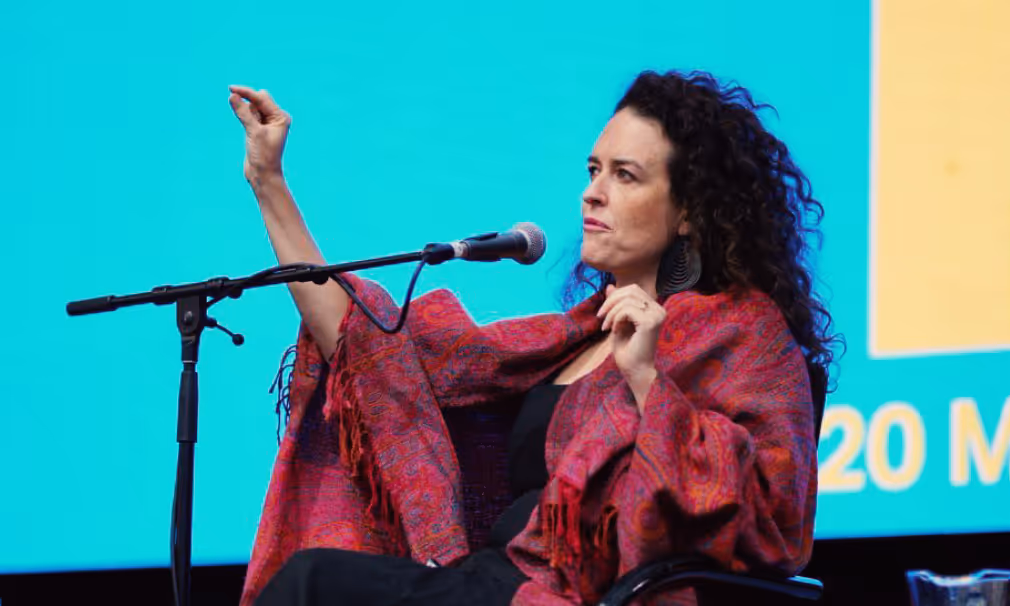 Woman with curly hair speaking into a microphone while gesturing with her hand, wearing a red patterned shawl.
