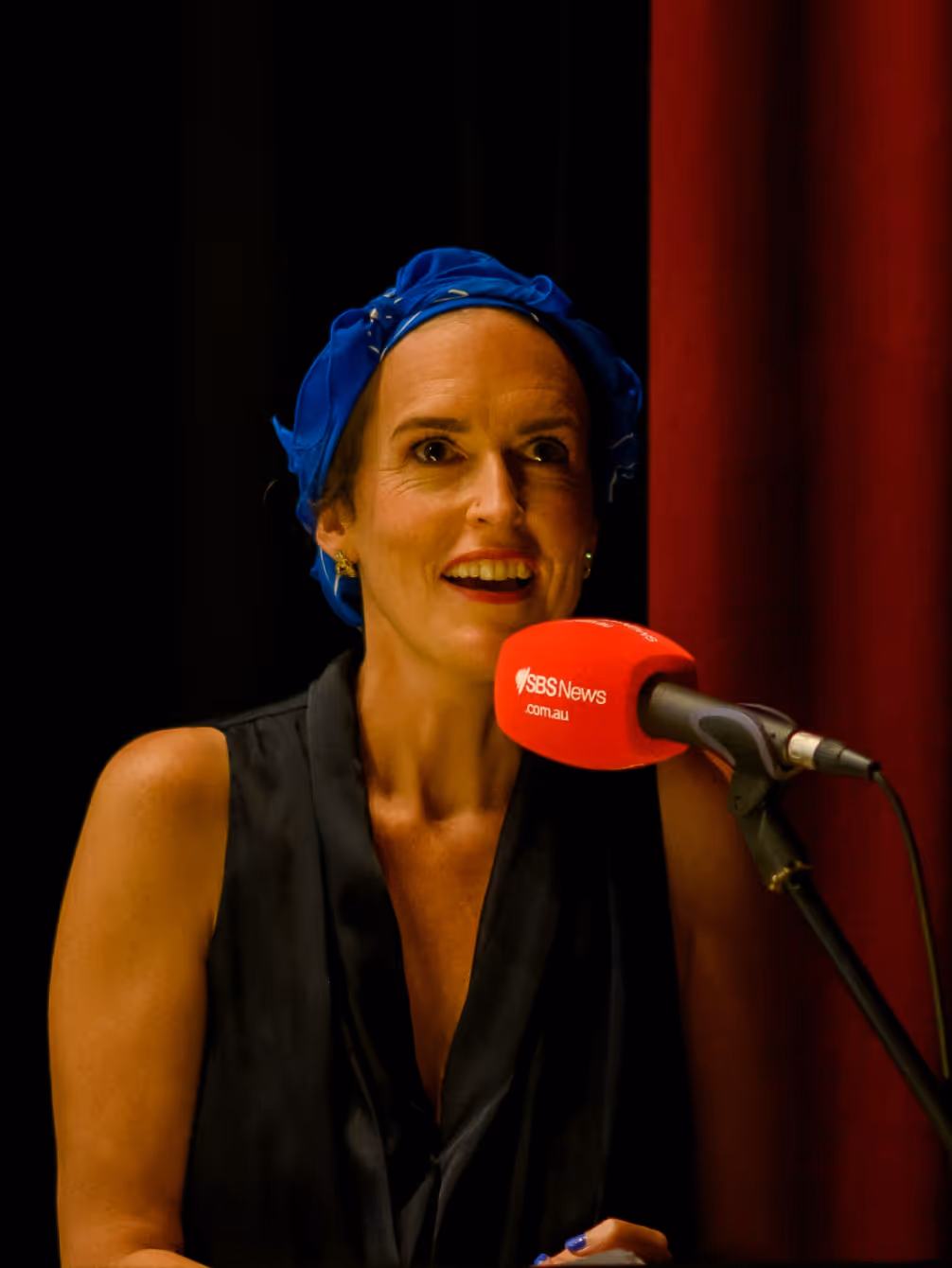 Woman wearing a blue headscarf and black sleeveless top speaking into a red SBS News microphone against a black and red curtain background.