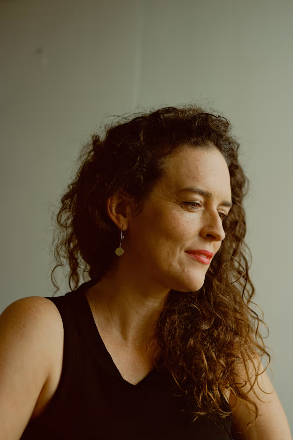 Woman with curly brown hair wearing a black sleeveless top, looking thoughtfully to the side against a plain background.