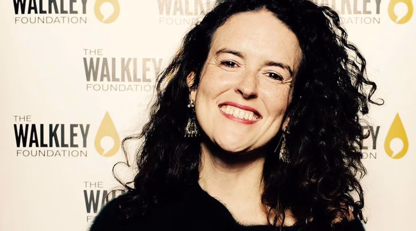 Smiling woman with curly dark hair wearing earrings in front of a Walkley Foundation backdrop.