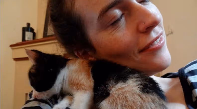 Woman smiling with eyes closed while holding a black, white, and orange calico cat on her shoulder indoors.