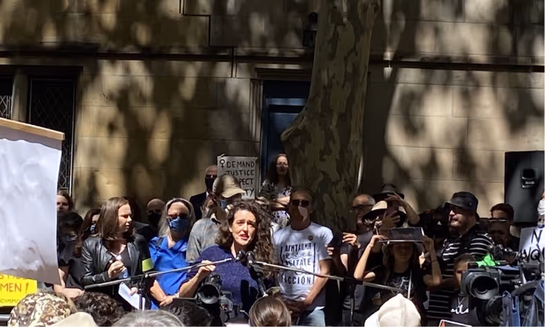 Woman with curly hair speaking into microphone at outdoor rally with crowd holding protest signs behind her.