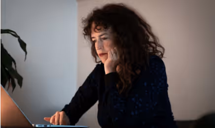 Woman with curly hair sitting at a desk, resting her chin on her hand while looking at a laptop.