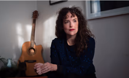 Woman with curly hair and red lipstick sitting indoors with an acoustic guitar in the background.